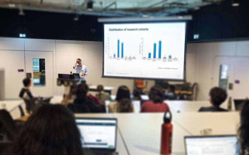 Students seated in a large classroom listening to a presenter, with laptops open and a presentation.